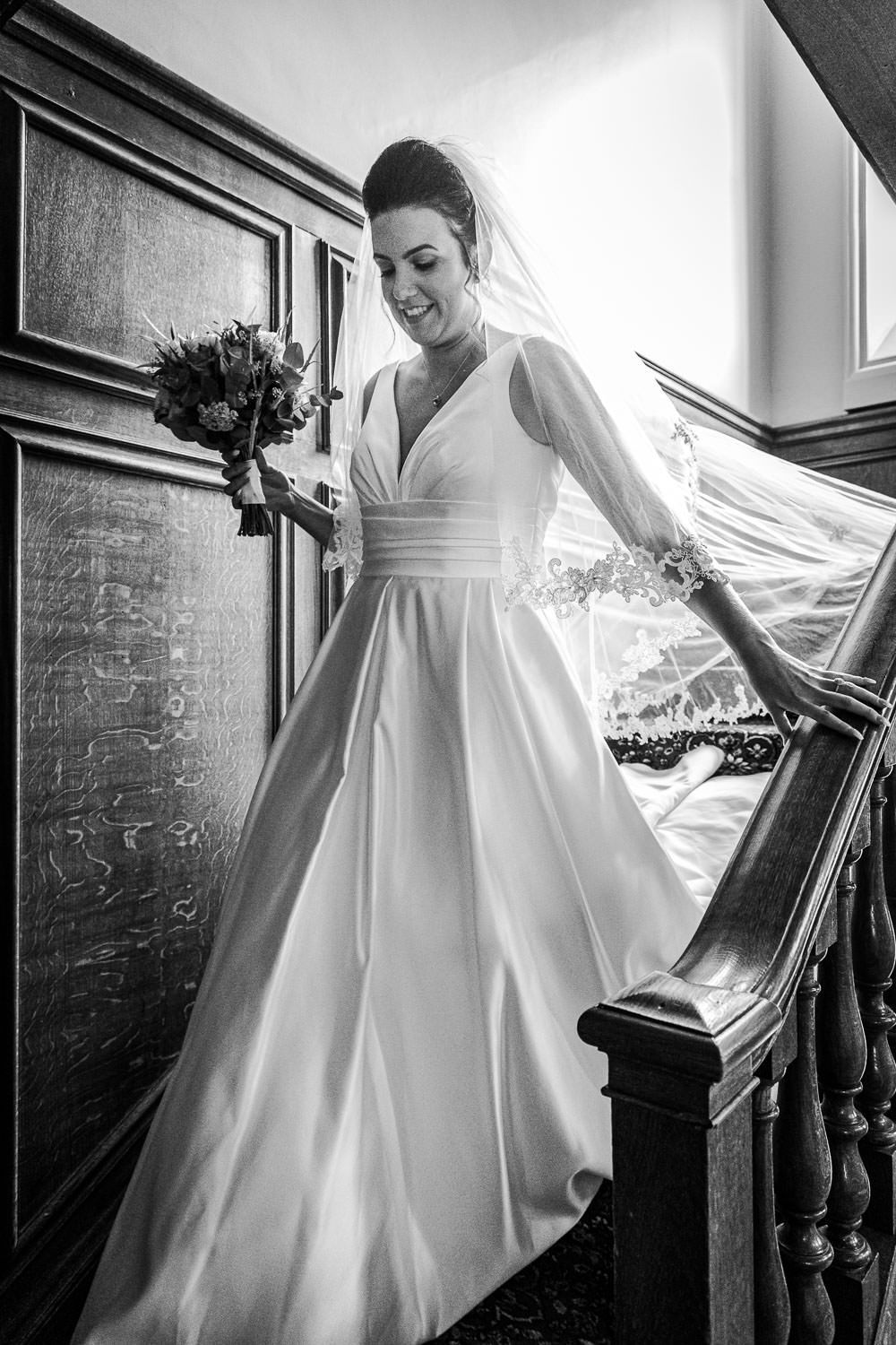 Elegant bride in white gown descending grand wooden staircase holding bouquet in black and white.