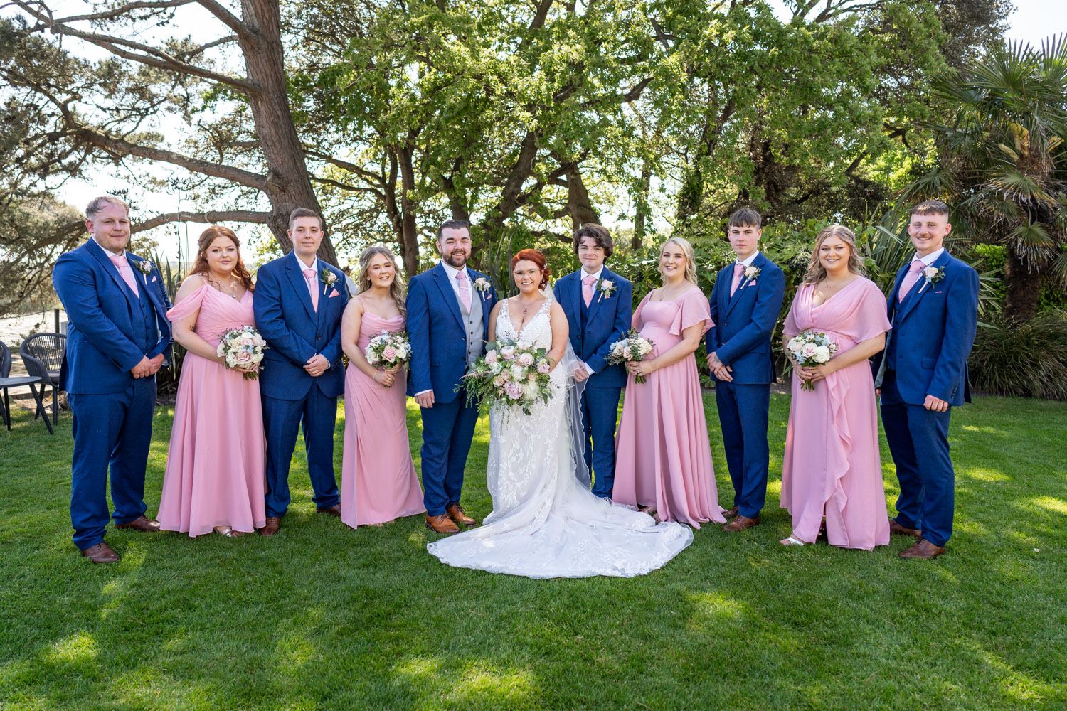 Wedding party in blue suits and pink bridesmaid dresses poses on green lawn during outdoor ceremony.