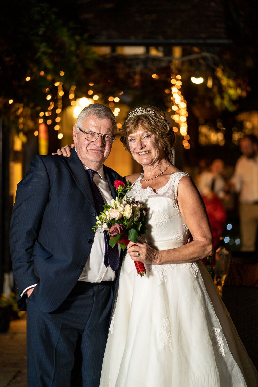 A couple celebrates their wedding day wearing formal attire with twinkling lights in the background.