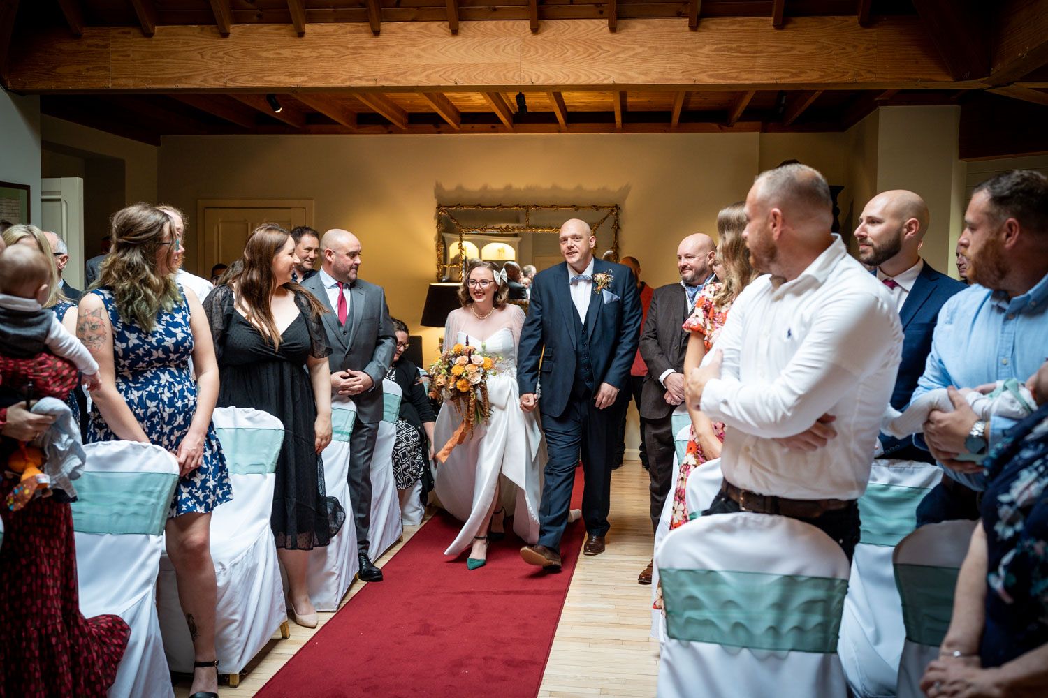 Wedding ceremony guests line the red carpet aisle inside a rustic wooden venue decorated with turquoise chair sashes.