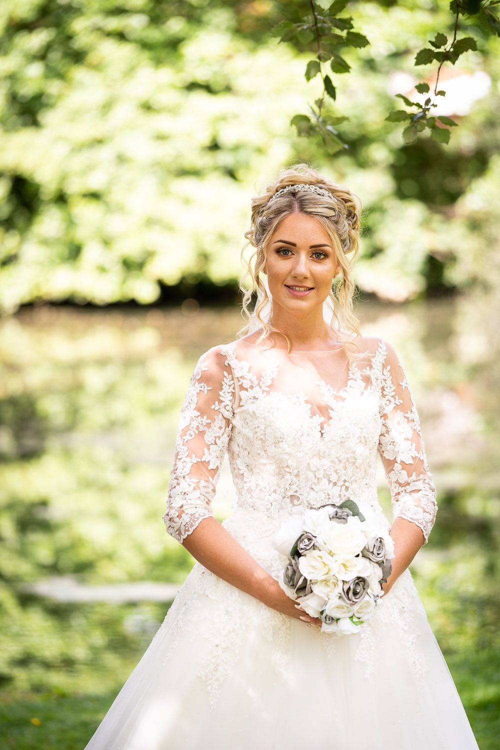 Elegant bride in lace wedding gown holding white bouquet, posing in lush green garden setting.