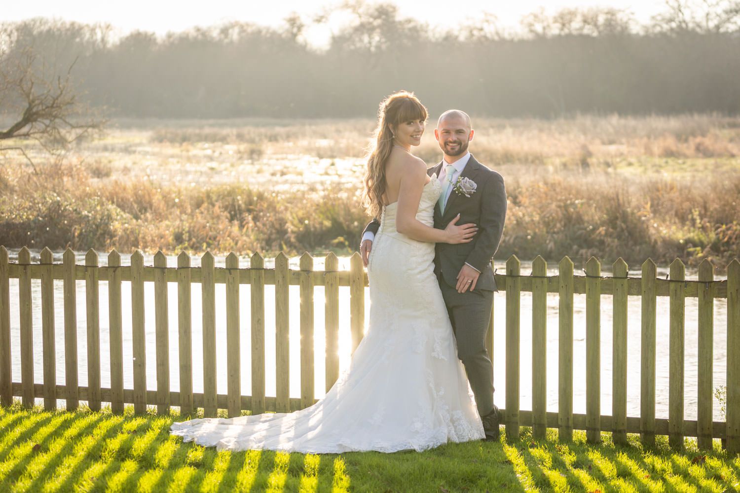 Bride and groom posing by a white picket fence in golden sunset light with a scenic meadow backdrop.