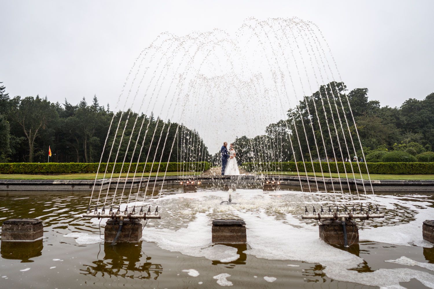 A dramatic water fountain display with white arching jets creating a tunnel effect over a reflective pool in a garden setting.