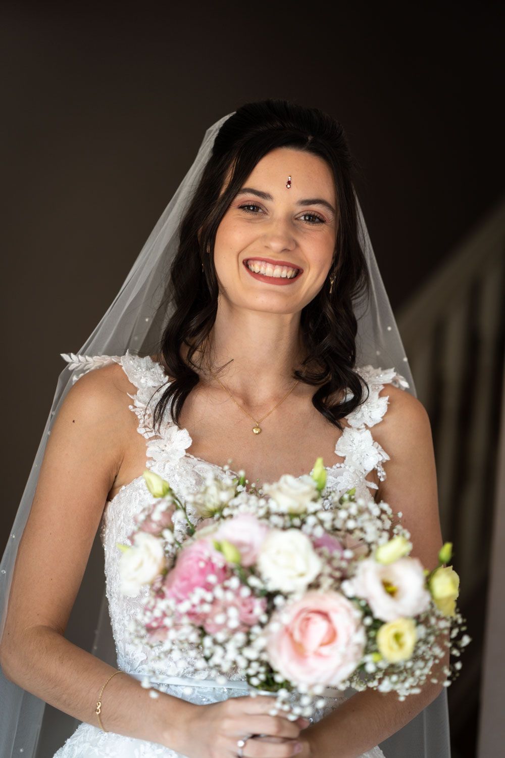Smiling bride in white lace dress holds delicate pink and white bouquet with baby's breath while wearing long veil.