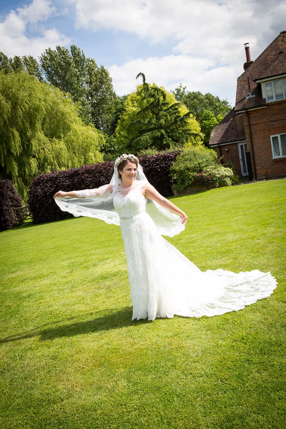A white wedding dress with flowing veil on display in a lush garden setting with weeping willows and hedges.