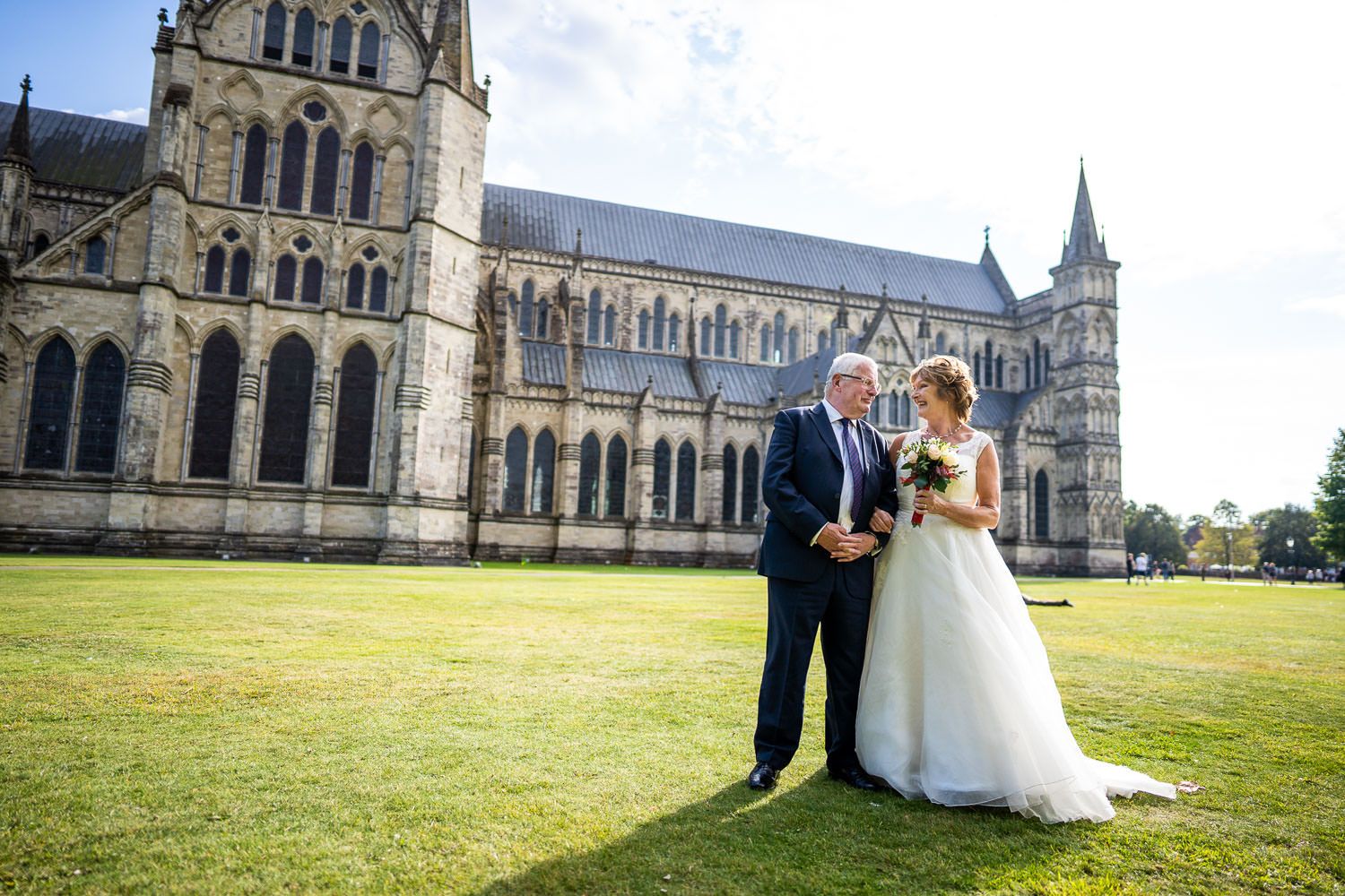 A couple in wedding attire poses on the green lawn in front of a majestic gothic cathedral on their special day.