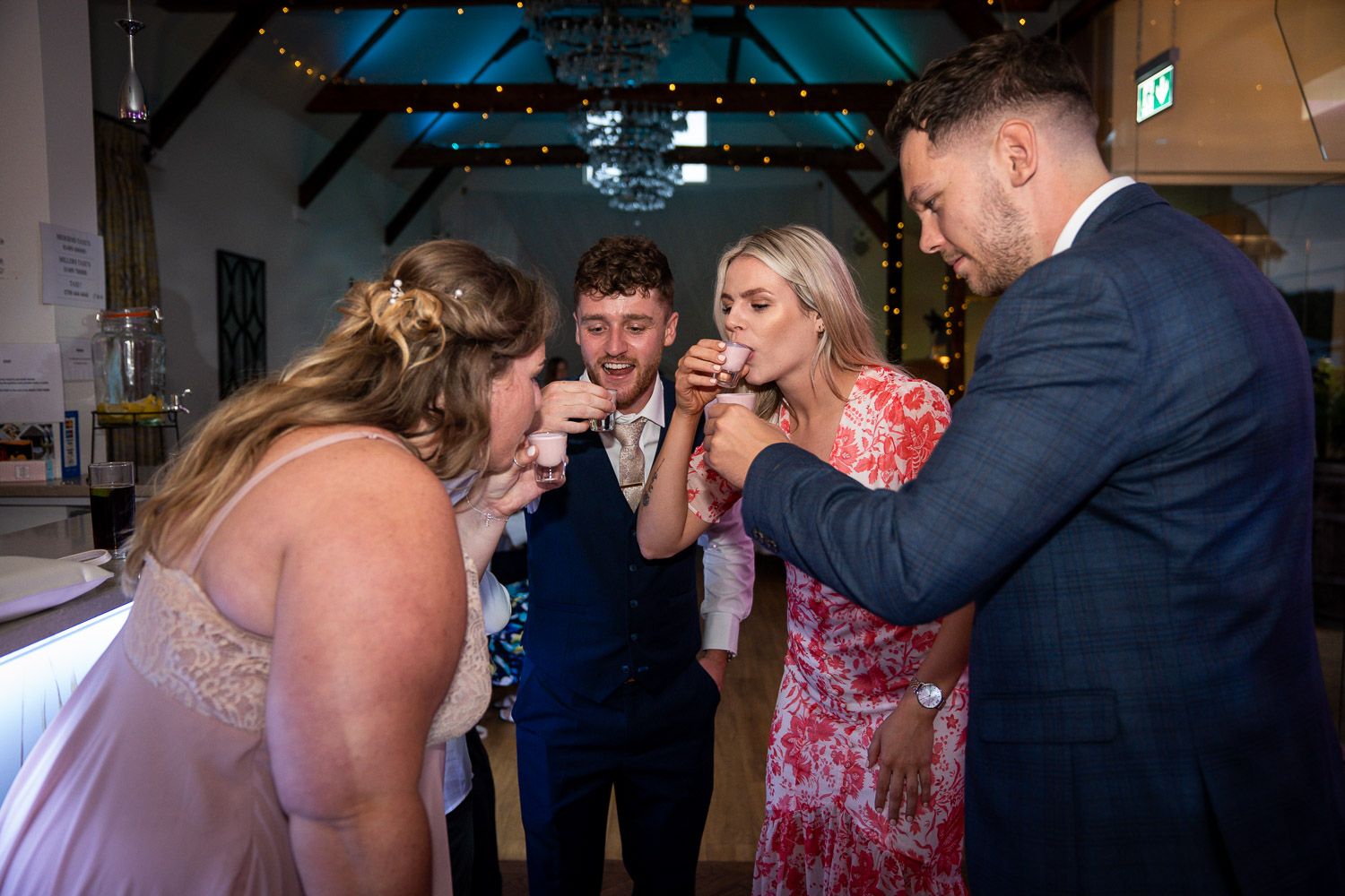 A group of wedding guests take shots together at a party reception with blue lighting in the background.