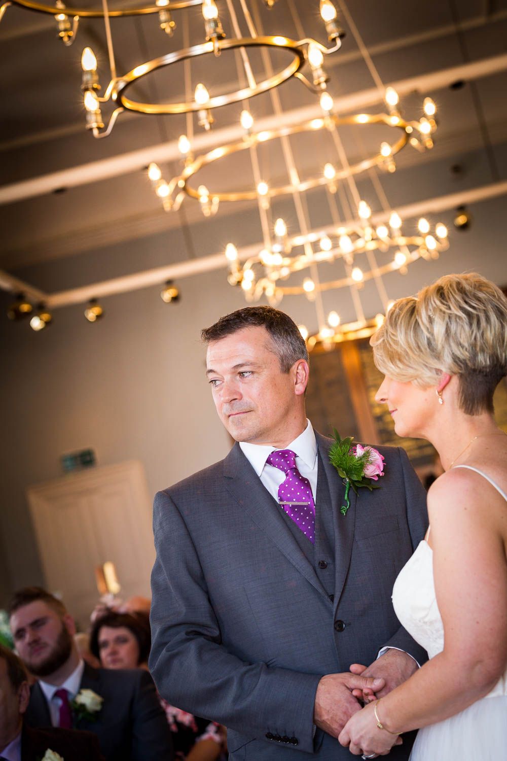 Wedding ceremony indoors with elegant circular chandeliers glowing warmly overhead during a special moment.