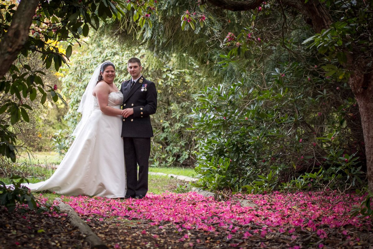 A couple stands together in a garden walkway covered with vibrant pink flower petals on the ground.