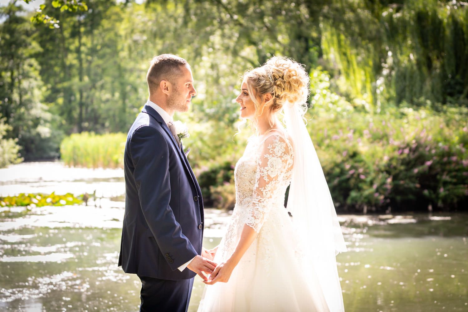 Bride and groom holding hands by a sunlit pond surrounded by lush green trees on wedding day.