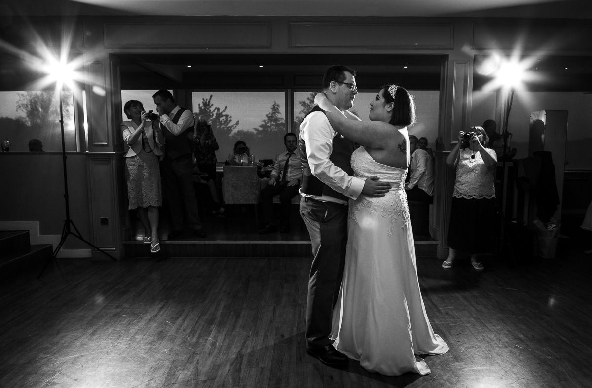 A couple shares their first dance on a dimly lit dance floor while photographers capture the romantic moment in black and white.
