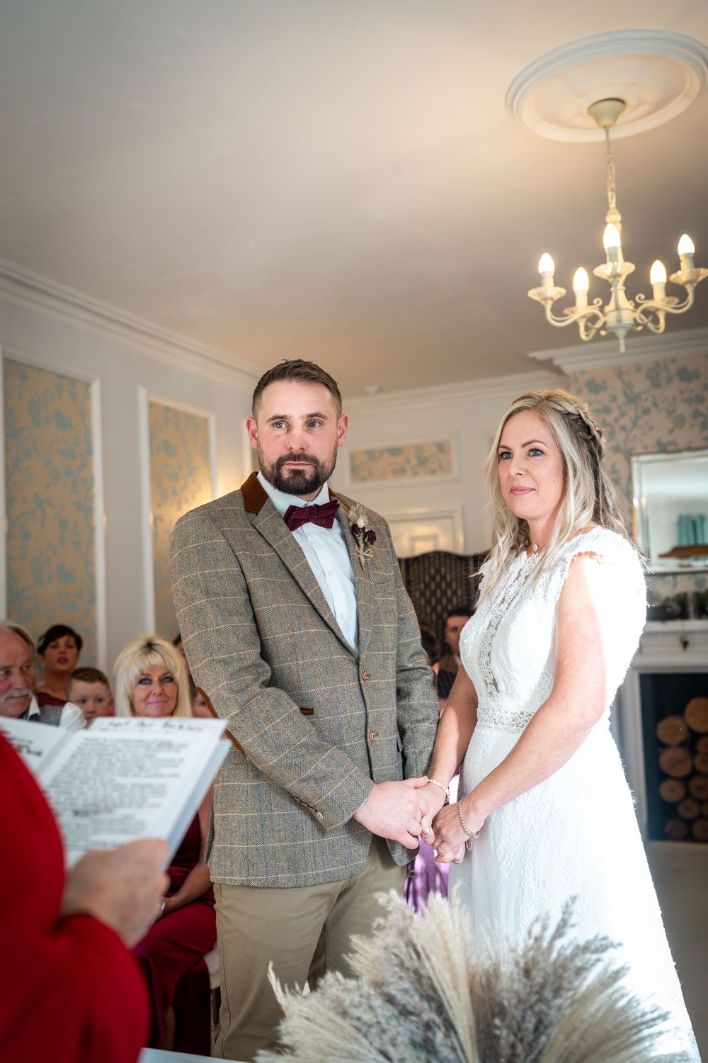 Indoor wedding ceremony with couple holding hands at the altar, decorated with a crystal chandelier and floral wall decor.