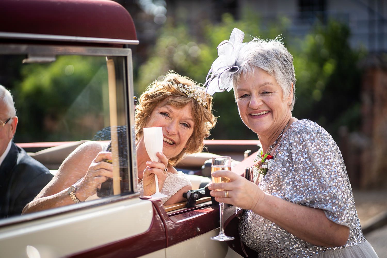 Two people toasting with wine glasses while sitting in a vintage car on a sunny day.