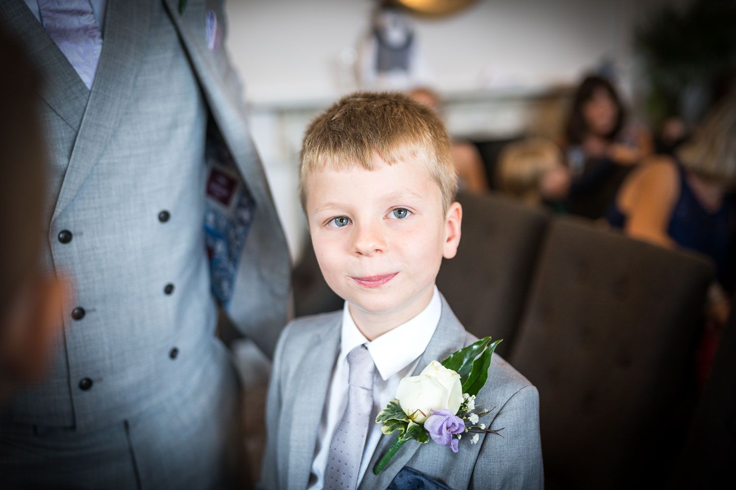 Young ring bearer in light gray suit and white boutonniere smiles during wedding celebration.