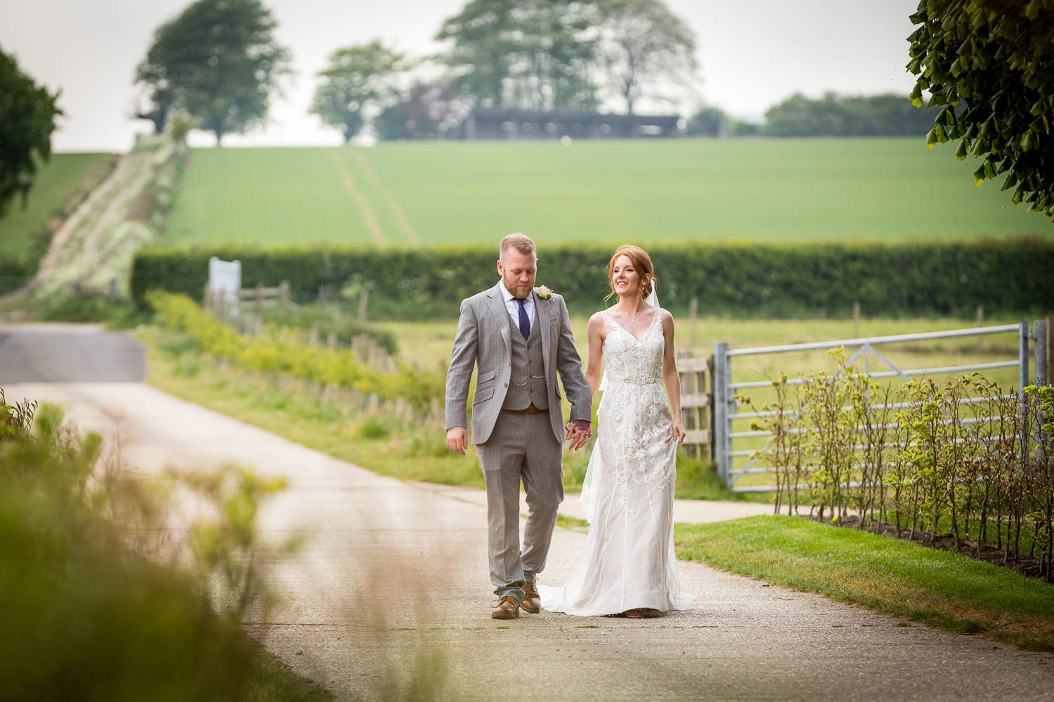 A couple in wedding attire walks hand in hand down a scenic country lane bordered by green fields and trees.