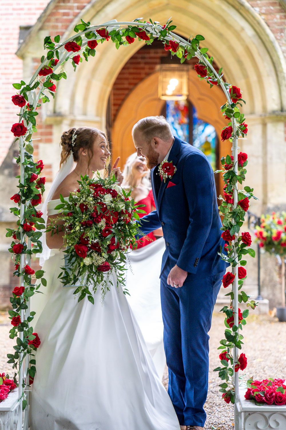 A wedding couple stands under a red rose-covered floral arch at a historic church entrance.