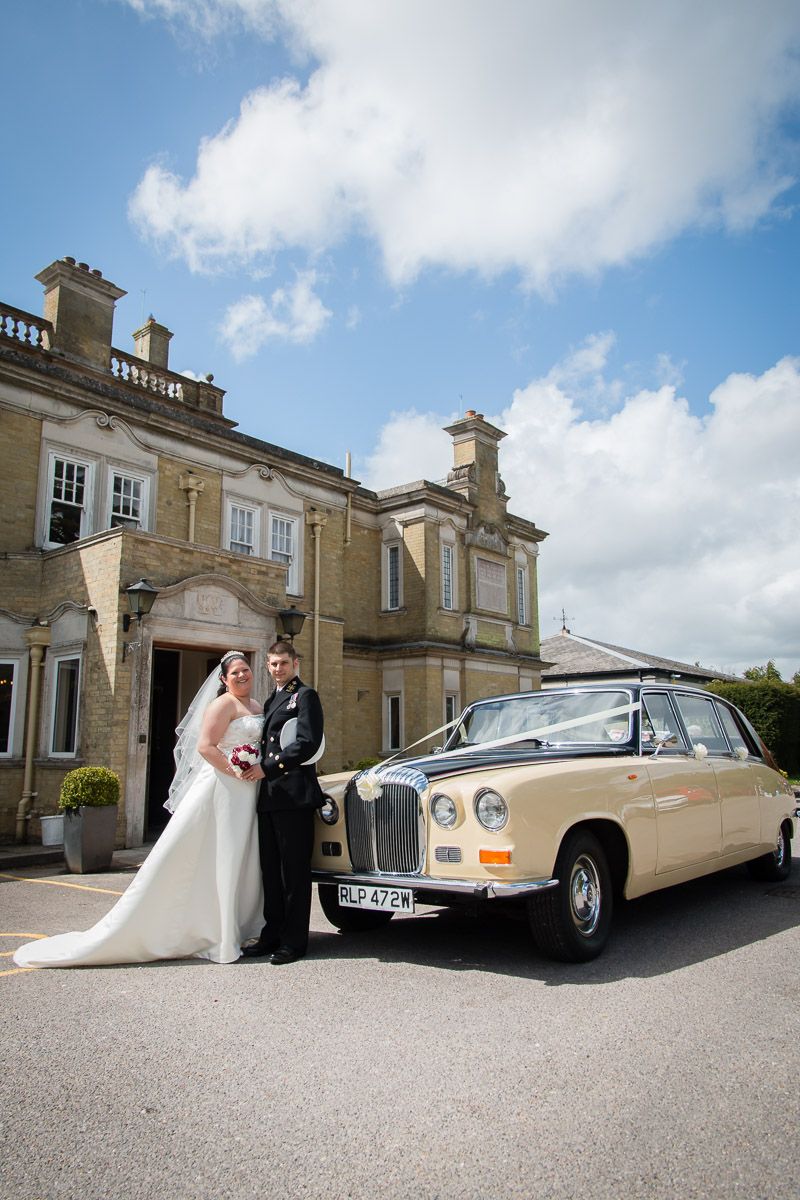 A classic cream Daimler limousine parked in front of an elegant stone mansion with wedding couple standing beside it.