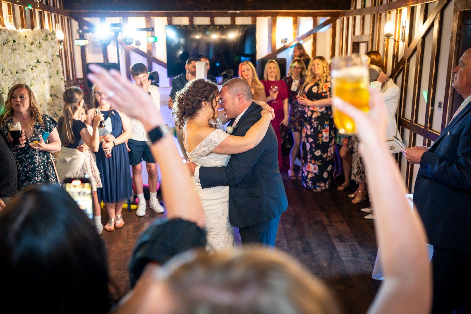 Dancing couple embraces while wedding guests raise toasts in rustic timber-framed venue.