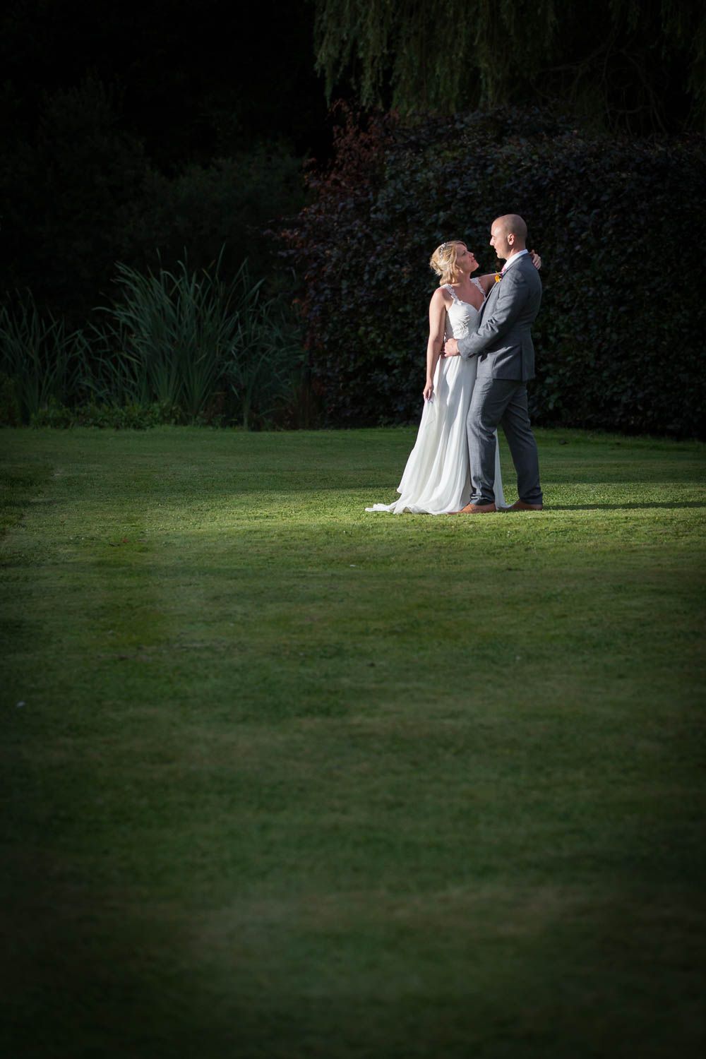 A wedding couple standing on an illuminated spot on a dark lawn with weeping willow trees in the background.