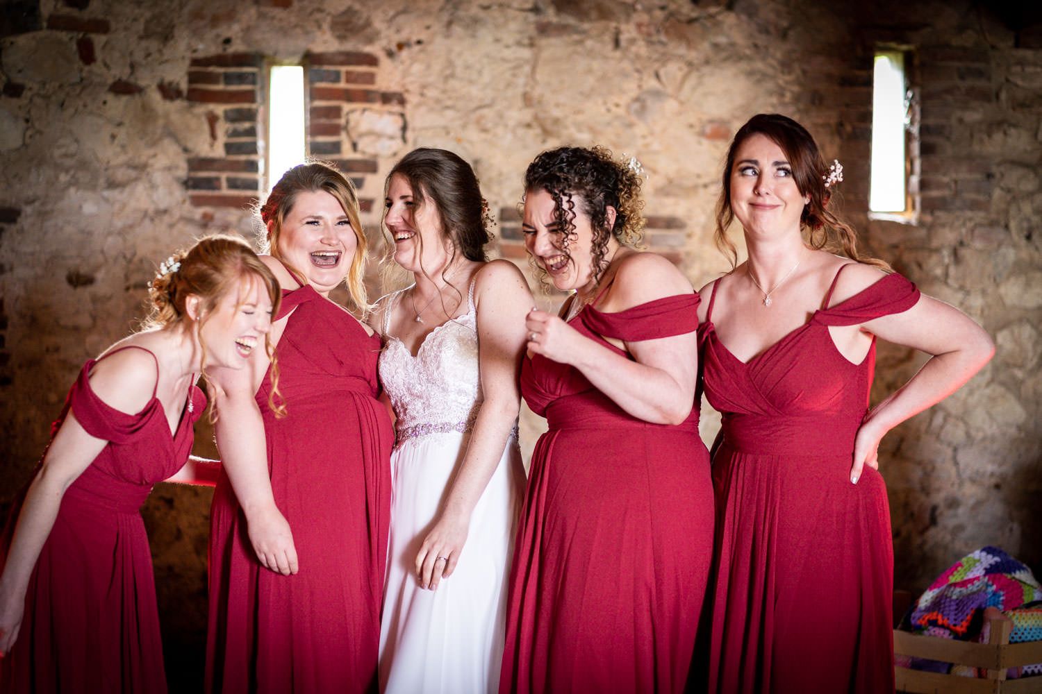 Bride laughing joyfully with four bridesmaids in red dresses inside a rustic stone venue.