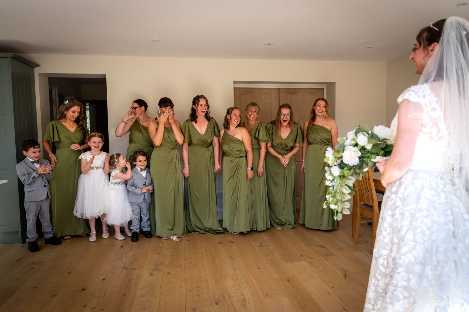 Bridesmaids in olive green dresses react emotionally as bride reveals her wedding gown indoors.