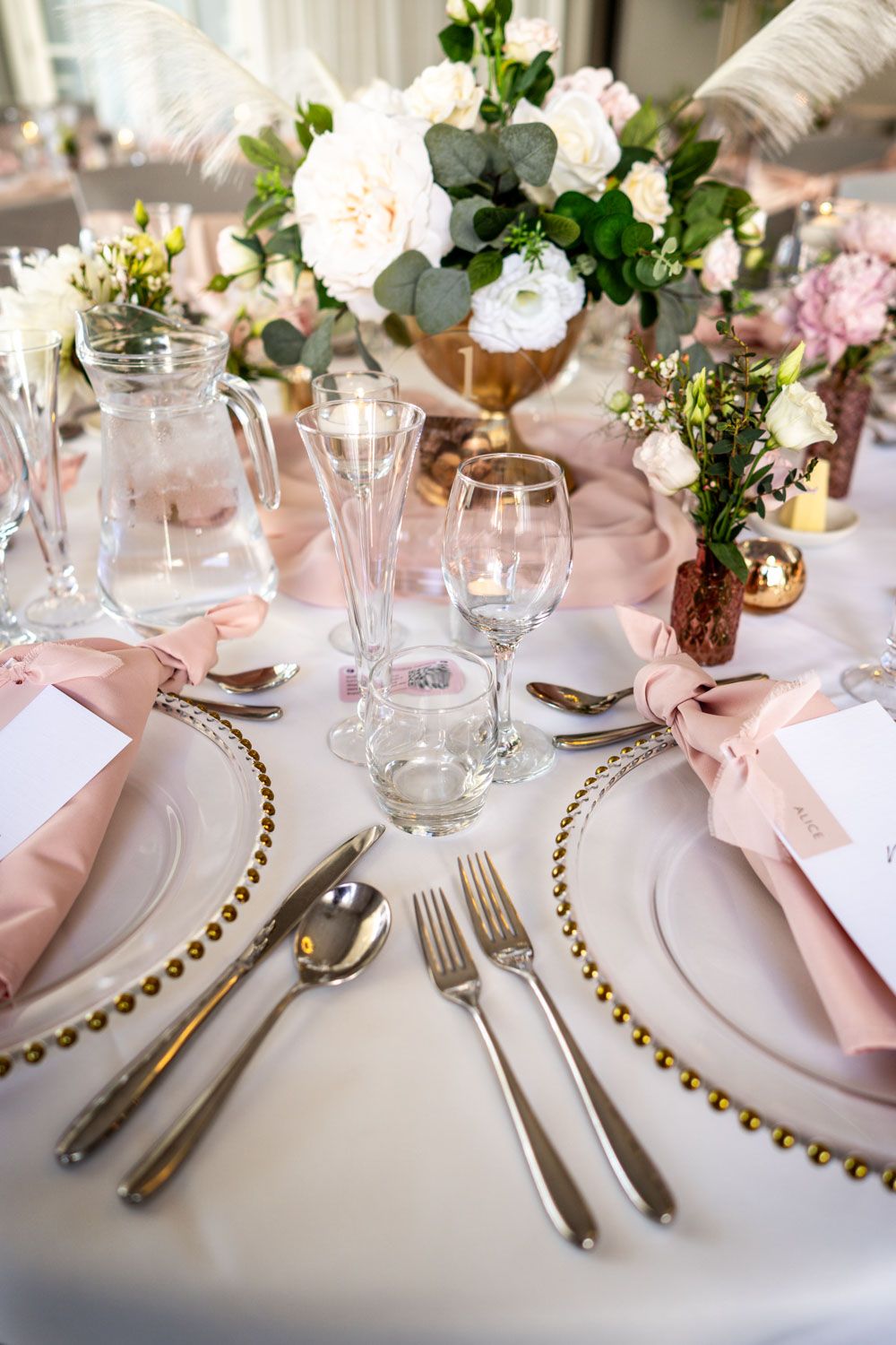 Elegant table setting with pink napkins, gold-rimmed plates, crystal glassware and floral centerpieces in metallic vases.