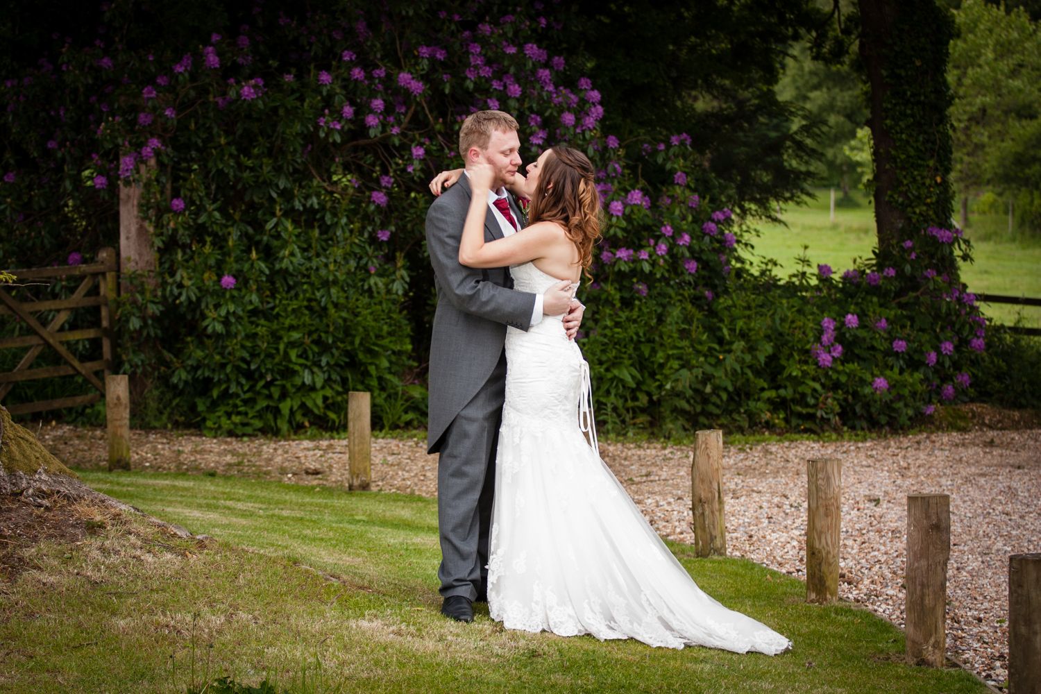 A romantic wedding couple embraces in a garden setting with blooming purple flowers and wooden fence posts.