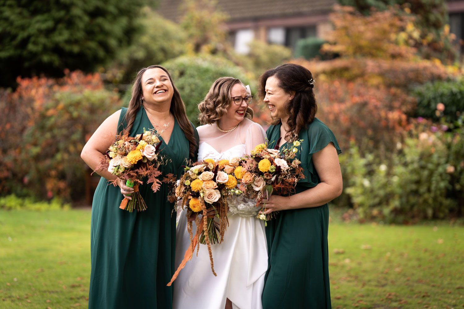 Three people in green bridesmaid dresses and white wedding gown share laughter while holding vibrant autumn bouquets outdoors.