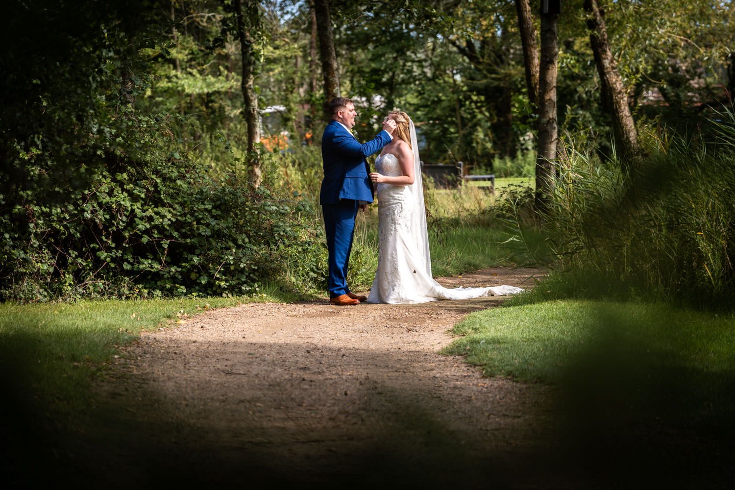 A couple in wedding attire embraces on a sunlit forest path surrounded by lush green trees and foliage.
