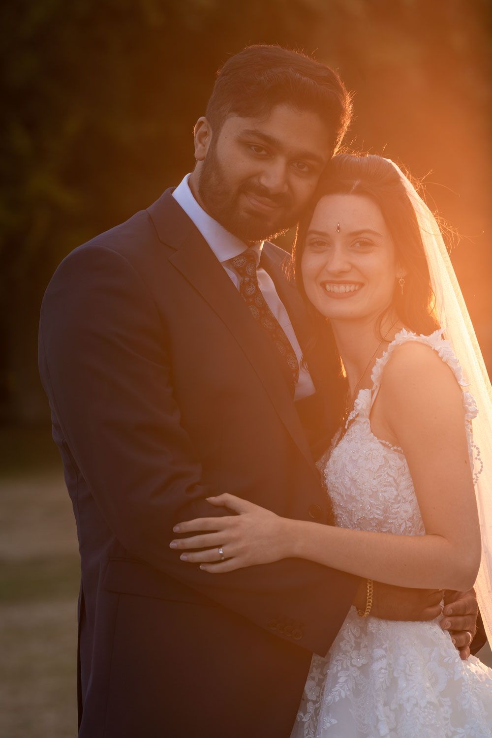 A happy couple embraces during their sunset wedding photoshoot, with bride in white lace gown and groom in dark suit.