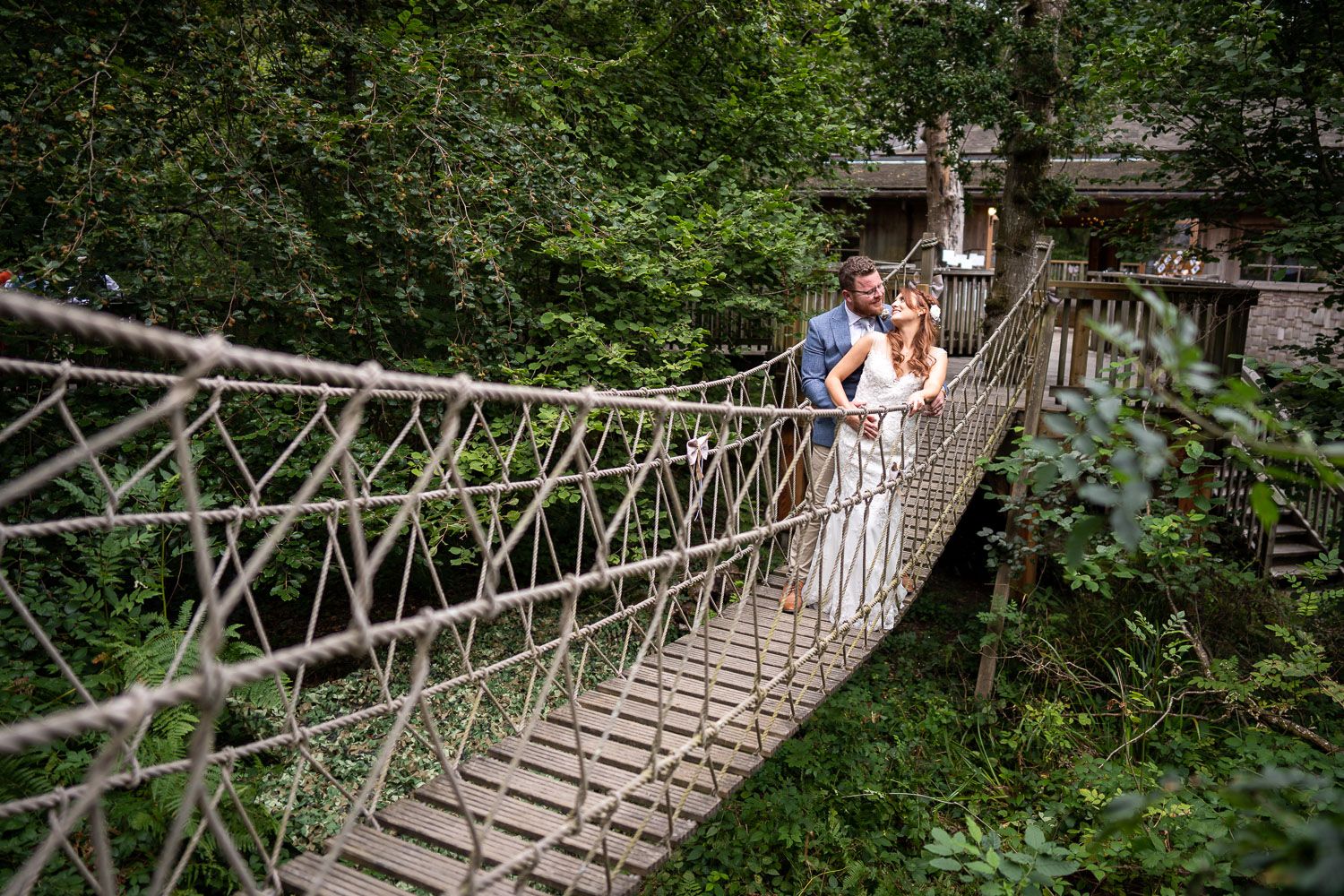 A couple embraces on a rustic wooden rope bridge surrounded by lush green forest scenery.