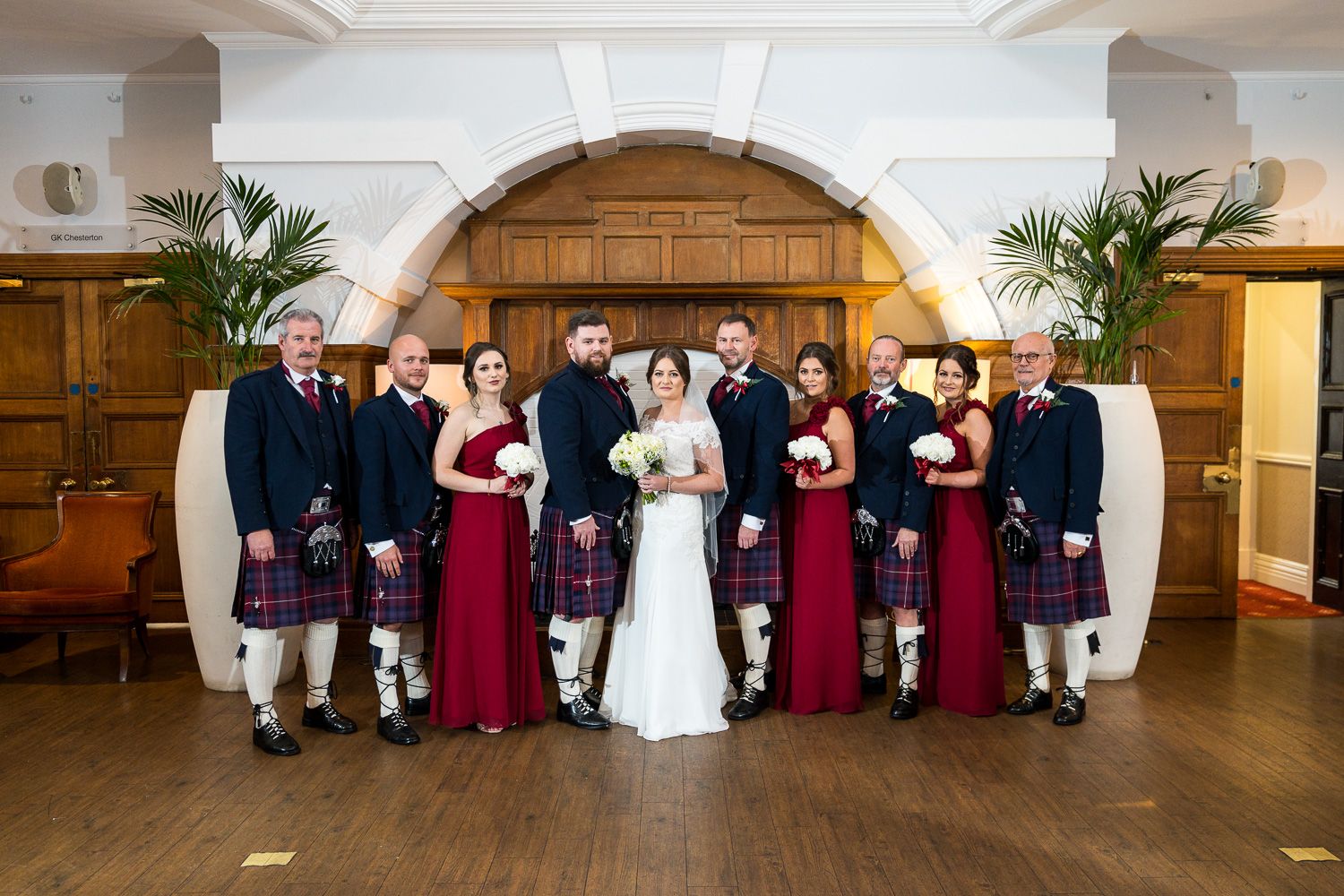Wedding party in burgundy dresses and kilts poses formally in an elegant room with white archways.