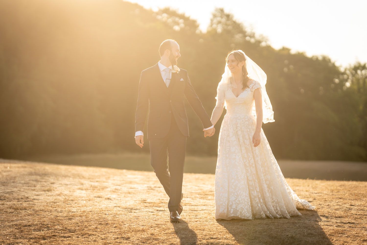 Bride and groom walking hand in hand across a sunlit field during golden hour at their outdoor wedding.