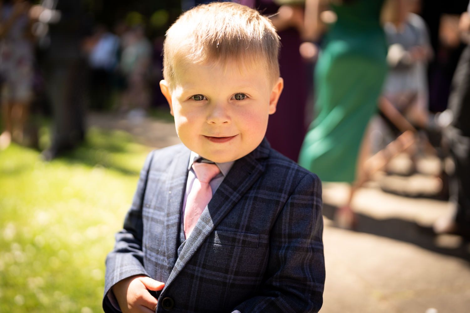 A young child in a plaid suit with a pink tie stands outdoors on a sunny day in a garden setting.