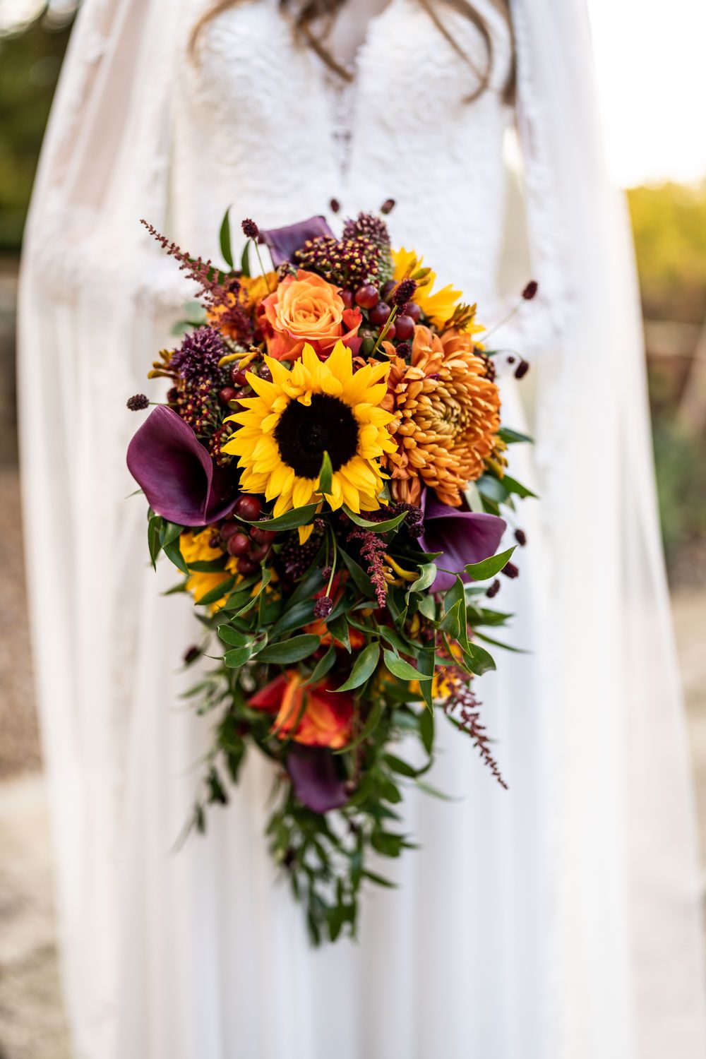 A cascading autumn bridal bouquet with sunflowers, purple calla lilies, and orange blooms against a white dress background.