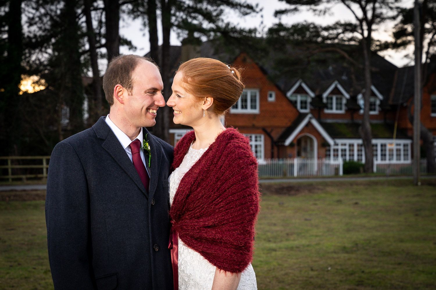A couple in formal attire stands together outdoors at dusk, with the bride wearing a red shawl and white dress.