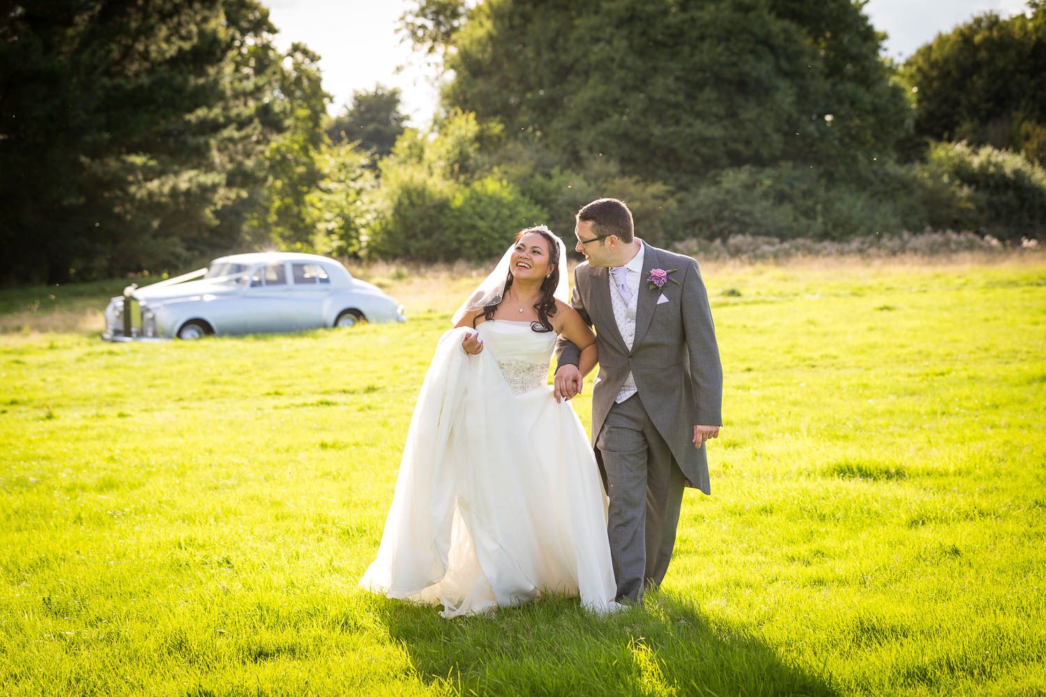 A couple in wedding attire walks together across a sunlit green meadow with a vintage car in the background.