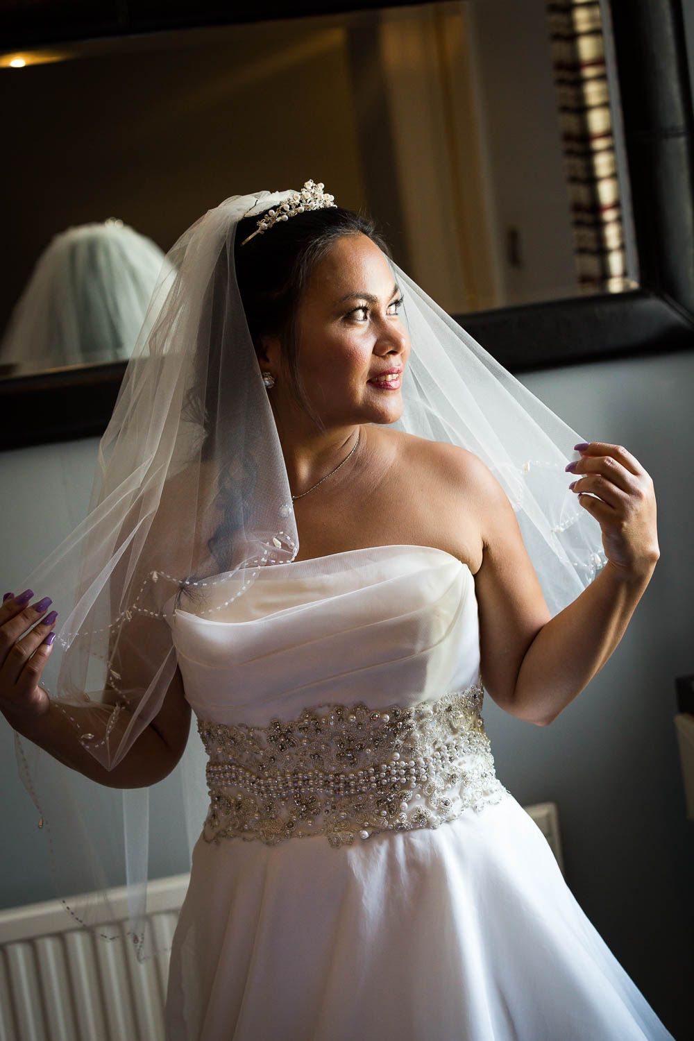 A bride in a white strapless wedding gown with beaded details and veil poses by a window in natural light.