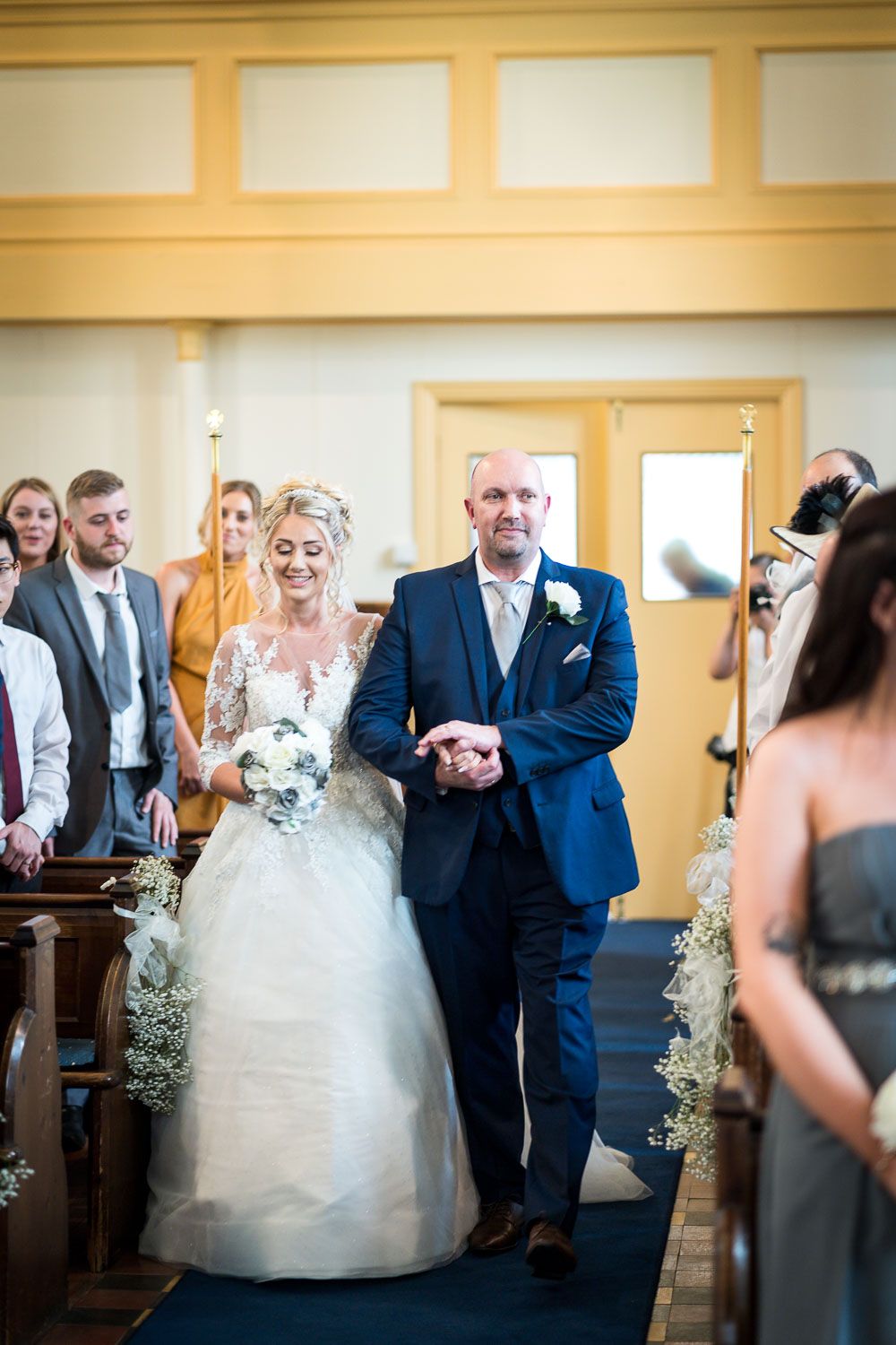 A bride wearing a white gown walks down the church aisle alongside a gentleman in a blue suit during a wedding ceremony.