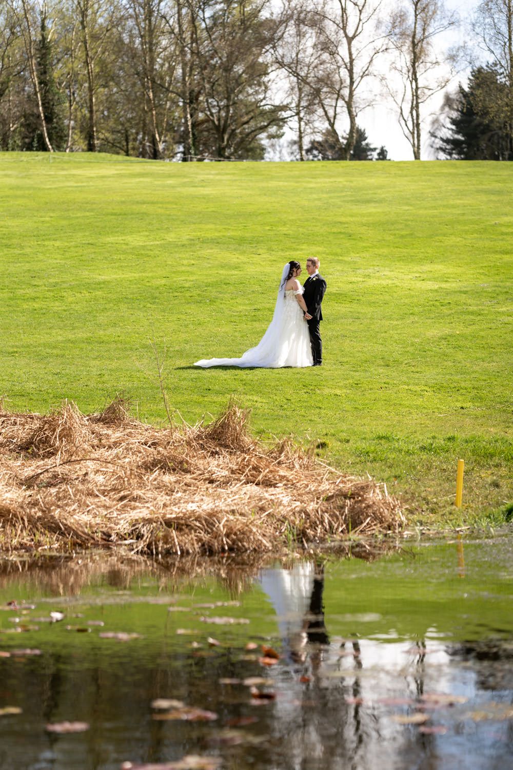 Newlyweds stand on lush green golf course hill beside a tranquil pond reflecting their silhouette on a sunny day.