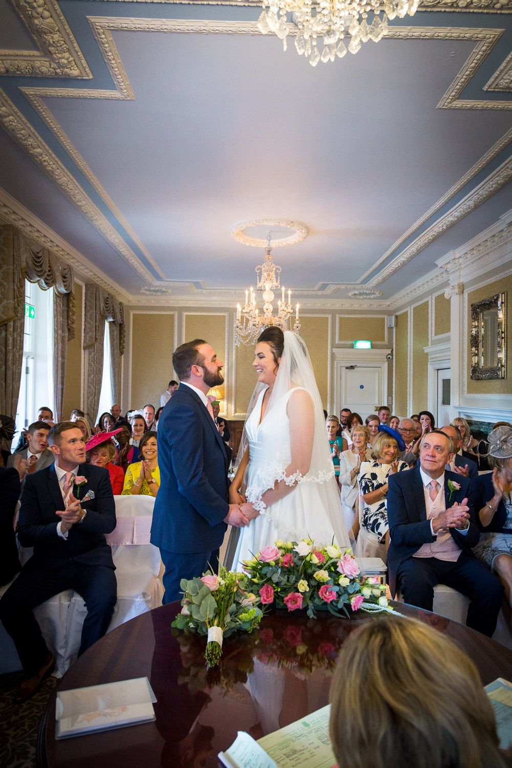 A wedding ceremony takes place in an elegant room with crystal chandeliers and ornate ceiling details in blue and gold.