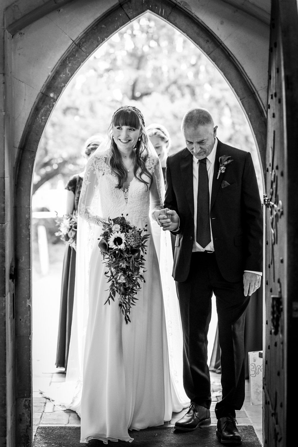 Black and white wedding photo of a couple walking through an arched doorway with the bride holding a sunflower bouquet.