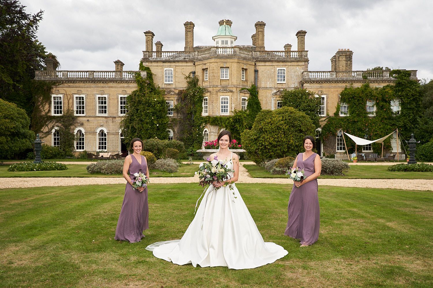 A wedding party poses on the lawn in front of a grand English manor house with classic Georgian architecture.