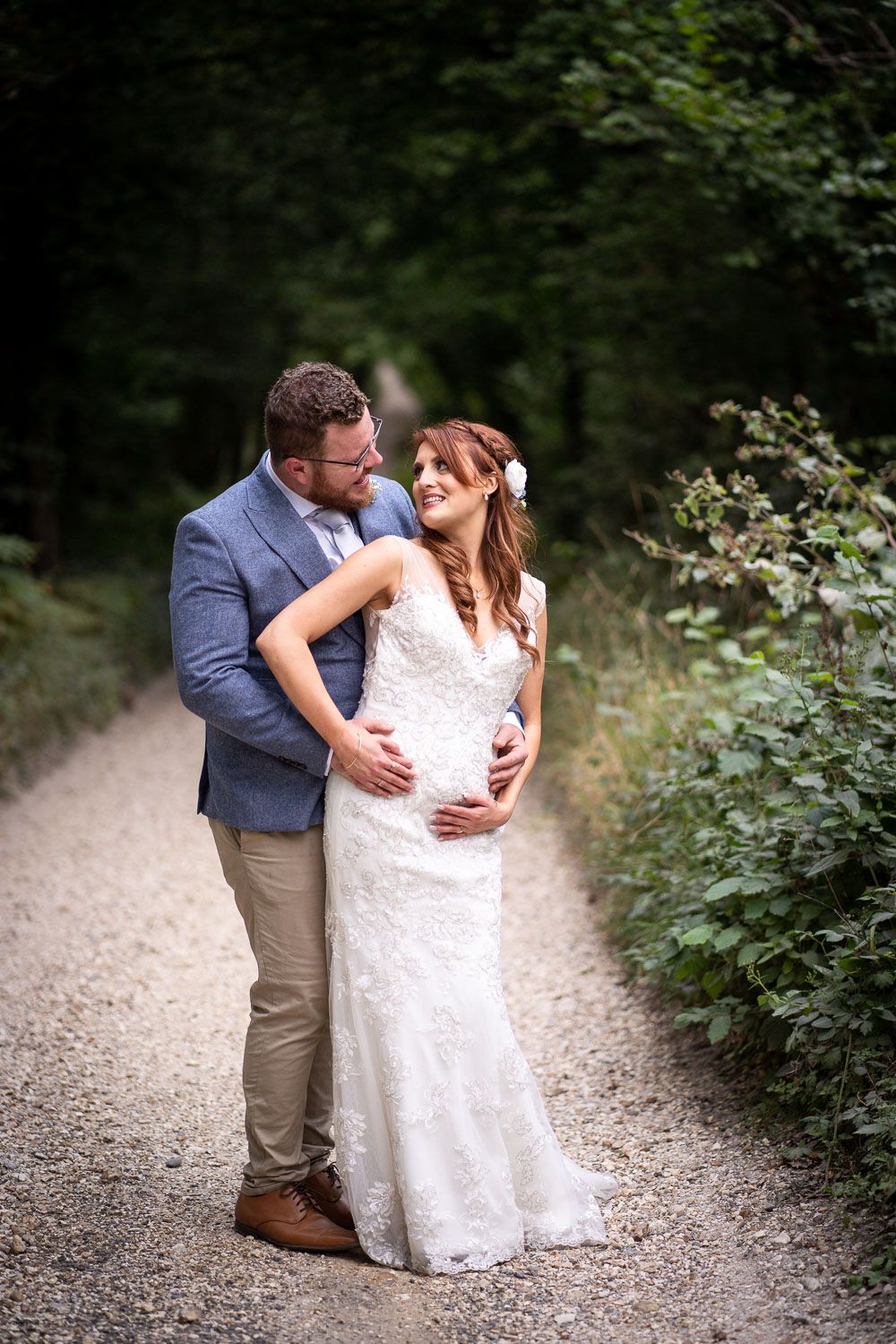 Couple sharing a romantic moment together on a gravel path surrounded by lush green foliage in formal wedding attire.