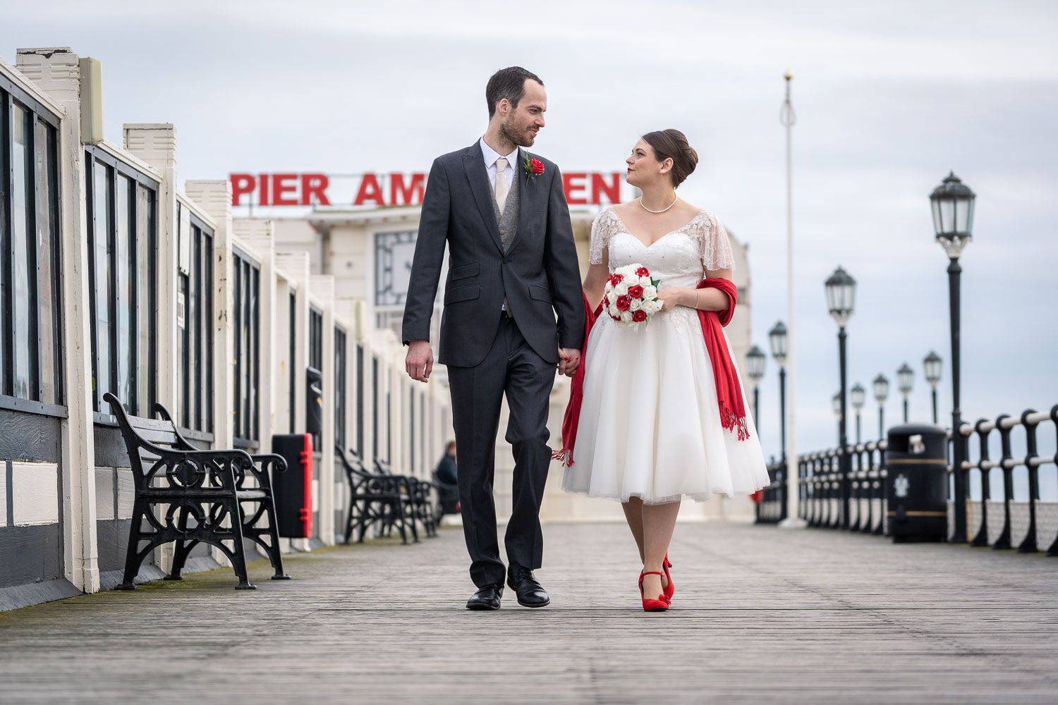 A couple in wedding attire walks together on a pier boardwalk with red accents and vintage lampposts visible.
