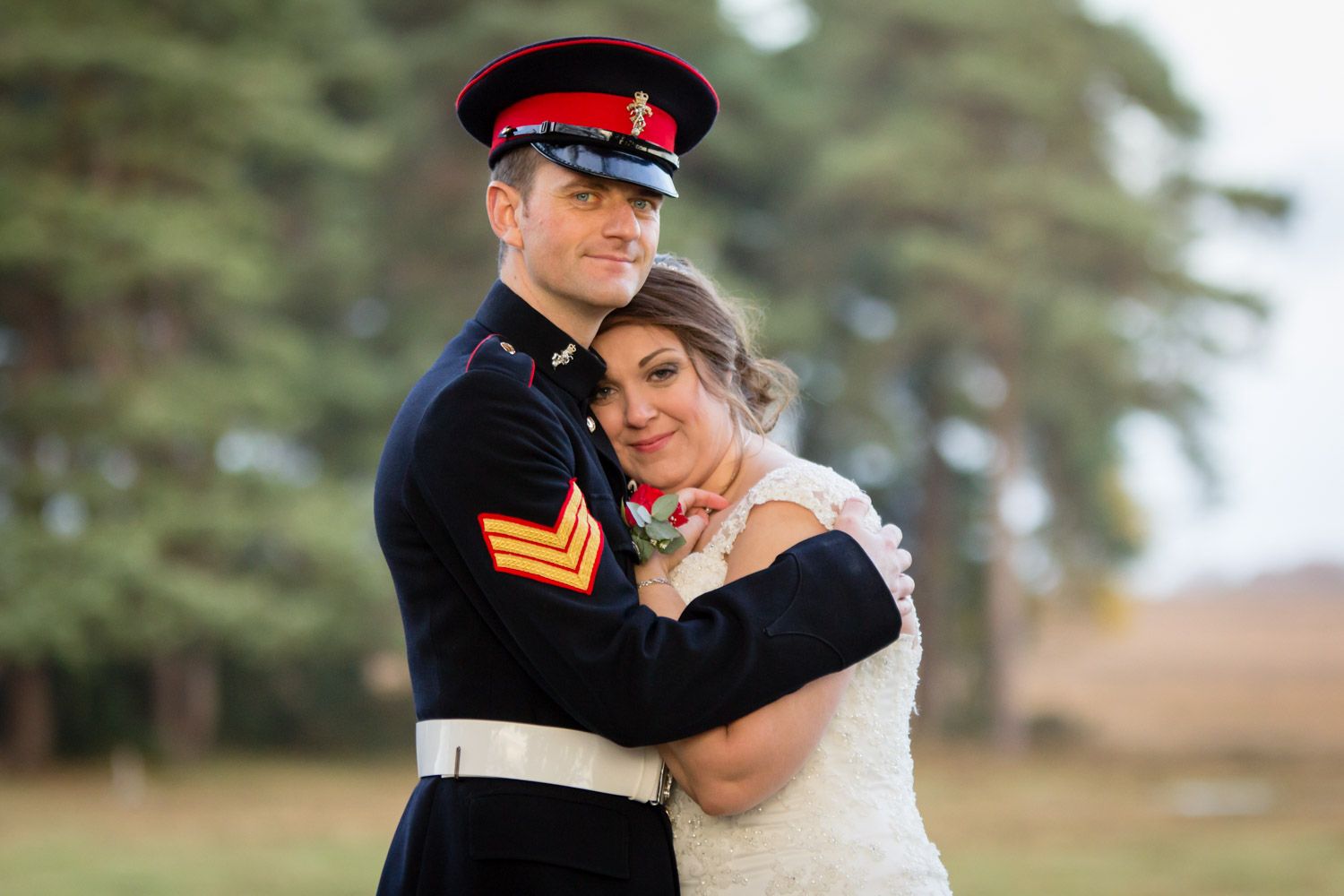 Military member in red and black dress uniform embraces their partner wearing a white gown during outdoor wedding photo.
