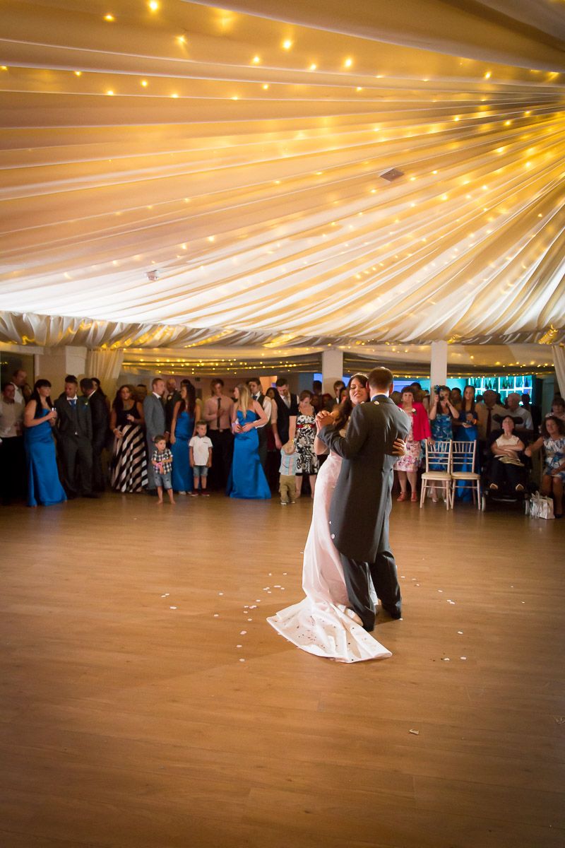 A couple shares their first dance under a beautifully draped tent ceiling with twinkling lights as wedding guests watch.