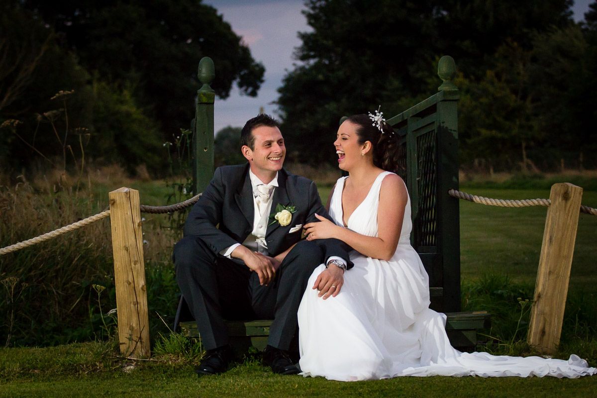 A bride and groom share a quiet moment while seated on a rustic wooden fence at dusk in a garden setting.
