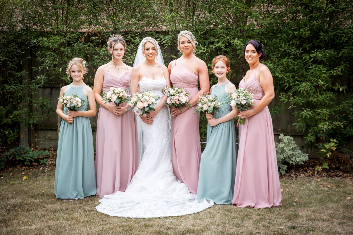 A wedding party poses outdoors wearing alternating pink and blue dresses with white floral bouquets.