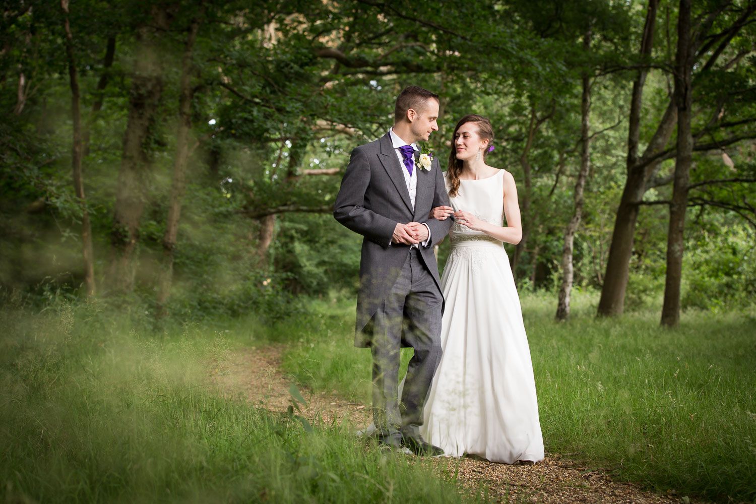 A newlywed couple walks together on a tree-lined path in a lush green forest setting.